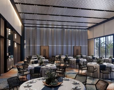 A grand, modern banquet hall with many round tables set for dining, featuring white tablecloths, blue runners, and floral centerpieces, under a textured, multi-paneled ceiling and large windows with views of palm trees.