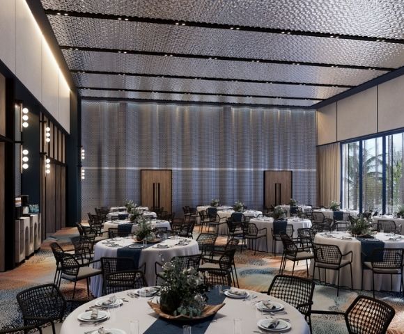 A grand, modern banquet hall with many round tables set for dining, featuring white tablecloths, blue runners, and floral centerpieces, under a textured, multi-paneled ceiling and large windows with views of palm trees.