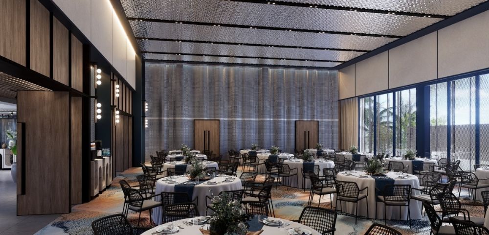 A grand, modern banquet hall with many round tables set for dining, featuring white tablecloths, blue runners, and floral centerpieces, under a textured, multi-paneled ceiling and large windows with views of palm trees.