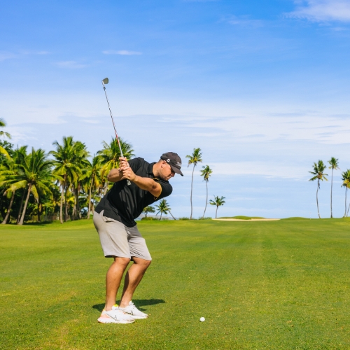 Golfer in black shirt swings an iron on a sunny tropical course, with palm trees, blue sky, green fairway, and a golf ball visible.