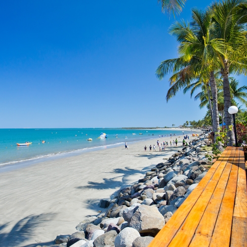 Sunny tropical beach with turquoise water, palm trees, swimmers, rocks, and a wooden boardwalk under a clear blue sky. Relaxing scene.