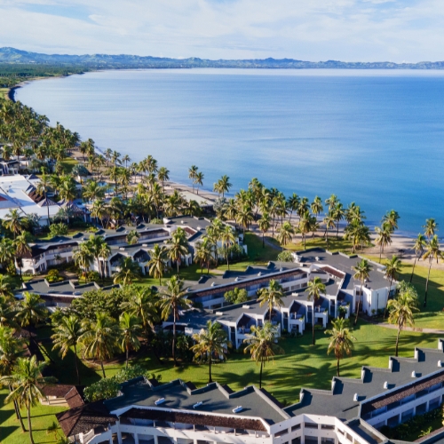 Aerial view of a tropical seaside resort with palm trees, white buildings, sandy beach, and a calm blue bay beneath a bright sky.