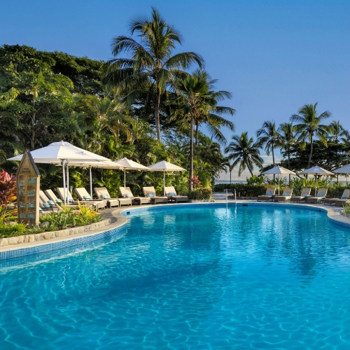 Serene tropical resort pool glistens beneath a clear blue sky, bordered by palm trees, white umbrellas, lounge chairs, and lush greenery.
