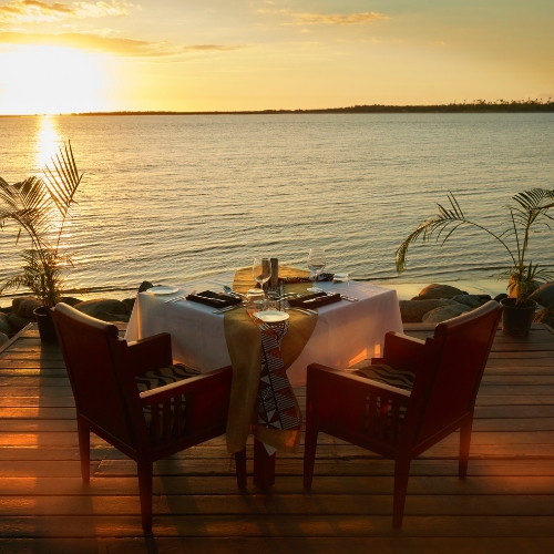 Romantic waterfront dinner table on a wooden deck at sunset, set for two with wine and candles, overlooking calm golden water and palms.