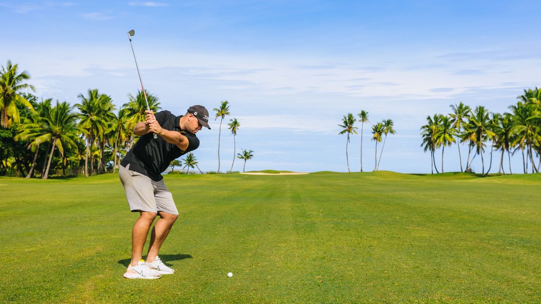 Golfer in black shirt swings an iron on a sunny tropical course, with palm trees, blue sky, green fairway, and a golf ball visible.