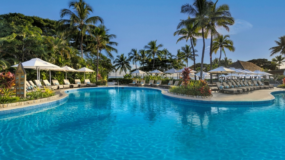 Serene tropical resort pool glistens beneath a clear blue sky, bordered by palm trees, white umbrellas, lounge chairs, and lush greenery.