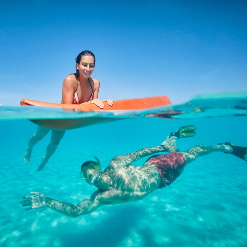 Split-level ocean scene: a smiling woman rests on a red paddleboard above, while a snorkeler swims below in clear turquoise water.