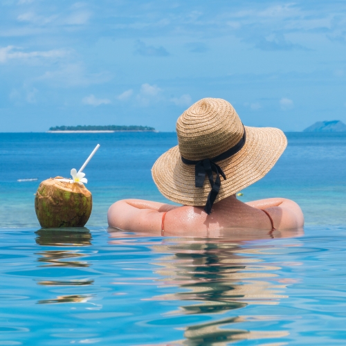 Back view of a woman in a straw sunhat relaxing in calm turquoise water beside a coconut drink, with islands and blue sky beyond.