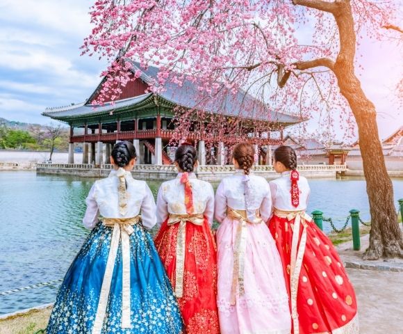 Four women in traditional Korean hanbok dresses stand by a serene body of water, with cherry blossom trees and a traditional pavilion in the background.
