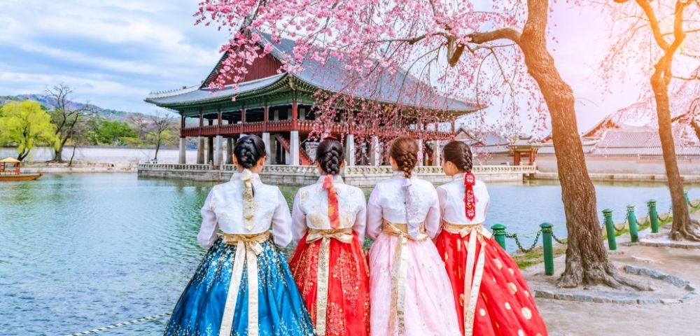 Four women in traditional Korean hanbok dresses stand by a serene body of water, with cherry blossom trees and a traditional pavilion in the background.