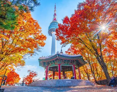A vibrant autumn scene featuring Namsan Tower in South Korea surrounded by colorful fall foliage, with a traditional pavilion in the foreground.