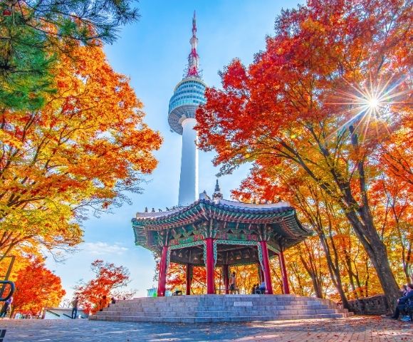 A vibrant autumn scene featuring Namsan Tower in South Korea surrounded by colorful fall foliage, with a traditional pavilion in the foreground.