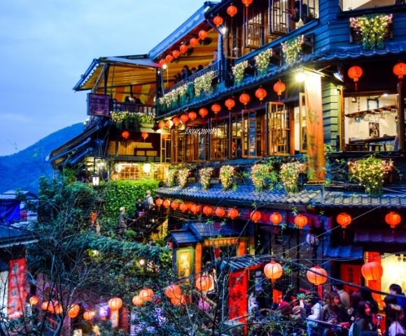 A picturesque street scene of a traditional Asian building adorned with red lanterns and fairy lights, set against a mountainous backdrop at twilight.