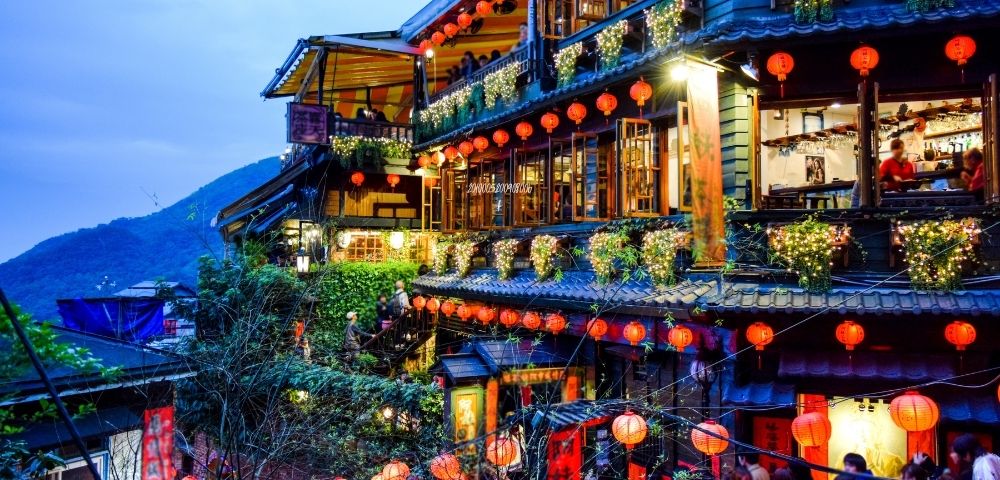 A picturesque street scene of a traditional Asian building adorned with red lanterns and fairy lights, set against a mountainous backdrop at twilight.