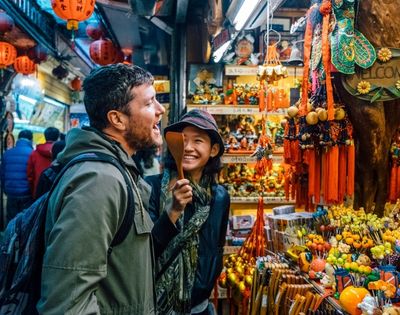 A man and a woman are laughing and interacting in a colorful market filled with various decorations and souvenirs.