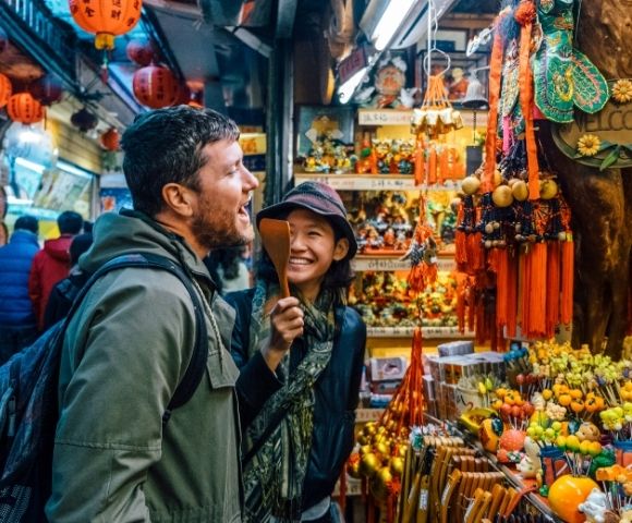 A man and a woman are laughing and interacting in a colorful market filled with various decorations and souvenirs.