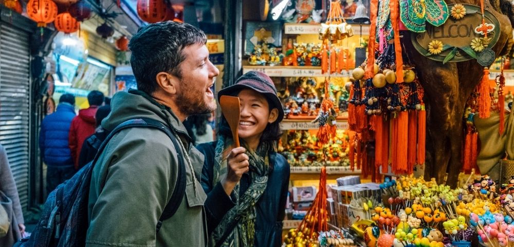 A man and a woman are laughing and interacting in a colorful market filled with various decorations and souvenirs.