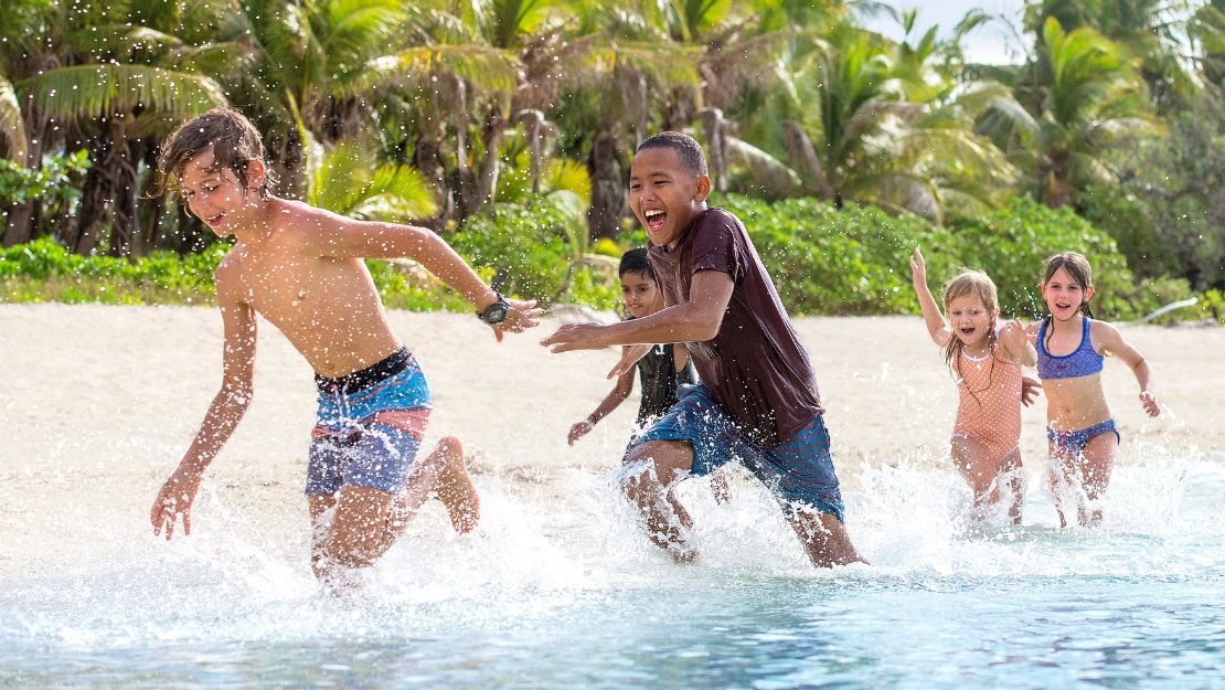 Five children laugh and run through shallow tropical water, splashing joyfully beneath bright sunlight and lush palm trees.