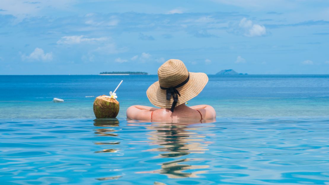 Back view of a woman in a straw sunhat relaxing in calm turquoise water beside a coconut drink, with islands and blue sky beyond.