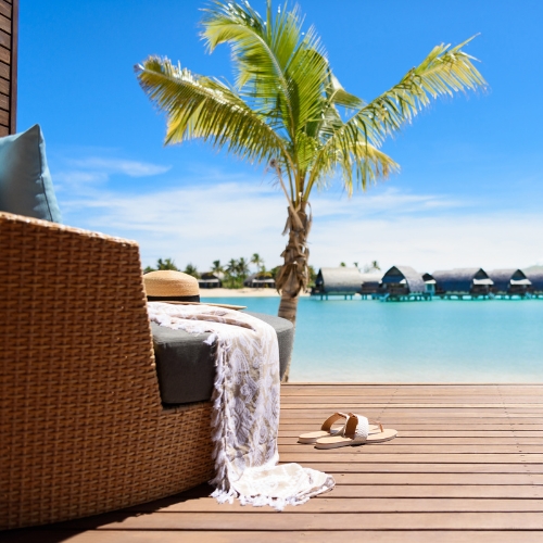 Sunny tropical resort deck with a palm tree, wicker lounge chair, white throw, and sandals overlooking a turquoise lagoon and bungalows.