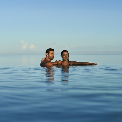Smiling couple relaxes chest-deep in a calm blue infinity pool, with the horizon blending into a clear sky, creating a peaceful mood.