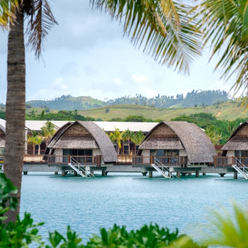 Tropical overwater bungalows with thatched roofs line a turquoise lagoon, framed by palms and green hills, creating a serene resort scene.