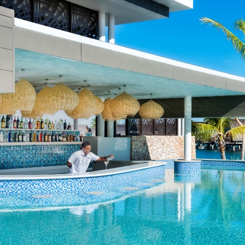 Poolside resort bar with bartender, woven pendant lamps, colorful bottles, turquoise water, palm trees, and bright tropical sunshine.