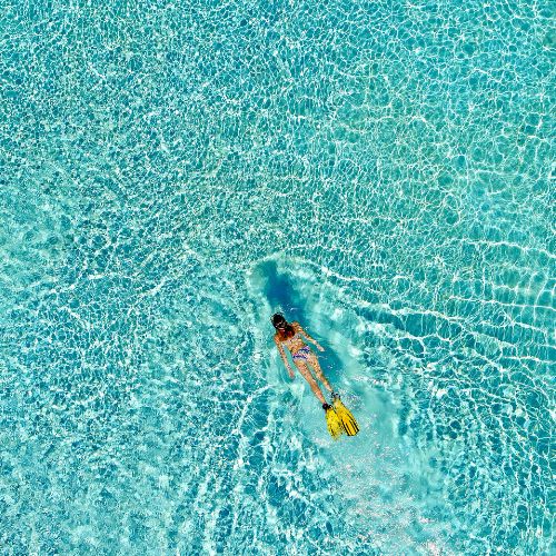 Aerial view of a swimmer wearing a mask and yellow fins gliding through bright turquoise pool water, creating ripples and a serene mood.