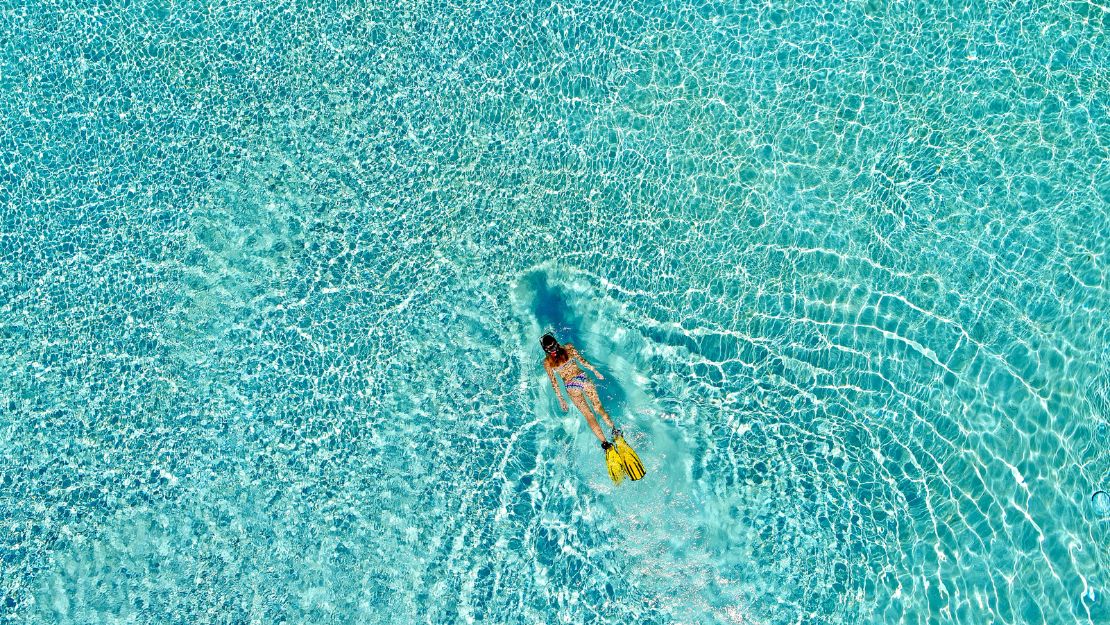 Aerial view of a swimmer wearing a mask and yellow fins gliding through bright turquoise pool water, creating ripples and a serene mood.