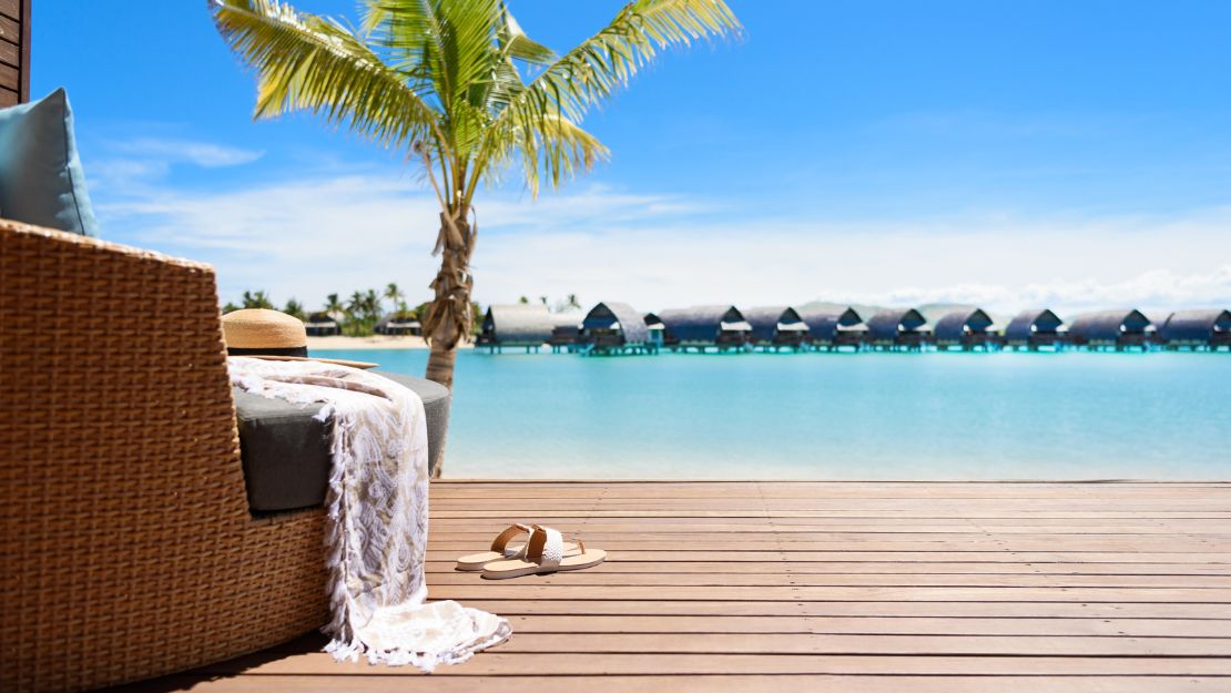Wicker lounge chair with white towel overlooks a turquoise lagoon, palm tree, wooden deck, sunhat, and overwater bungalows in serene sunlight.