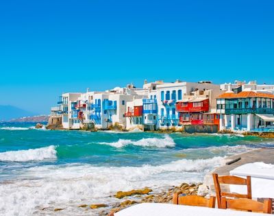 Bright seaside village with white and colorful buildings along a turquoise coast, foamy waves, rocky beach, and a calm blue sky.