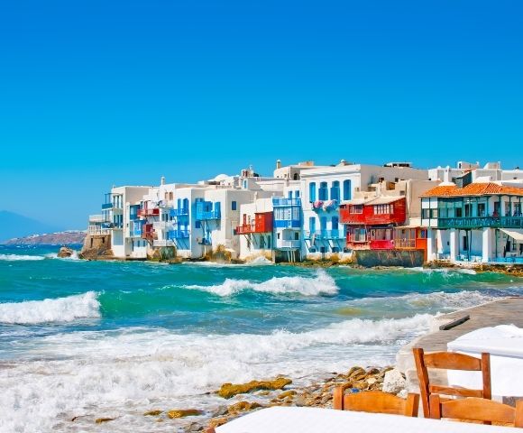 Bright seaside village with white and colorful buildings along a turquoise coast, foamy waves, rocky beach, and a calm blue sky.