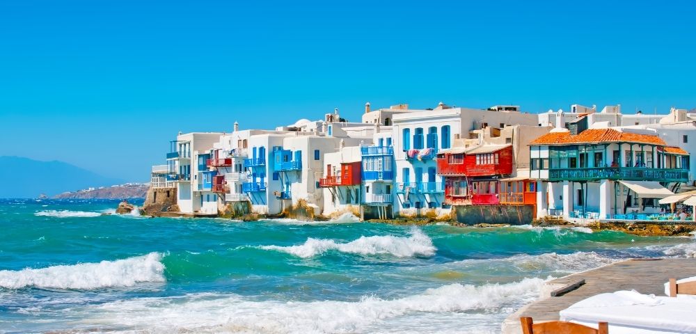 Bright seaside village with white and colorful buildings along a turquoise coast, foamy waves, rocky beach, and a calm blue sky.