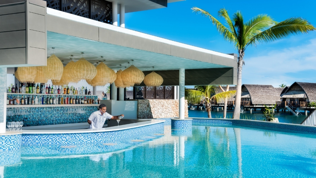Modern tropical resort swim-up bar with a bartender, woven pendant lights, turquoise pool, palm trees, and overwater bungalows.