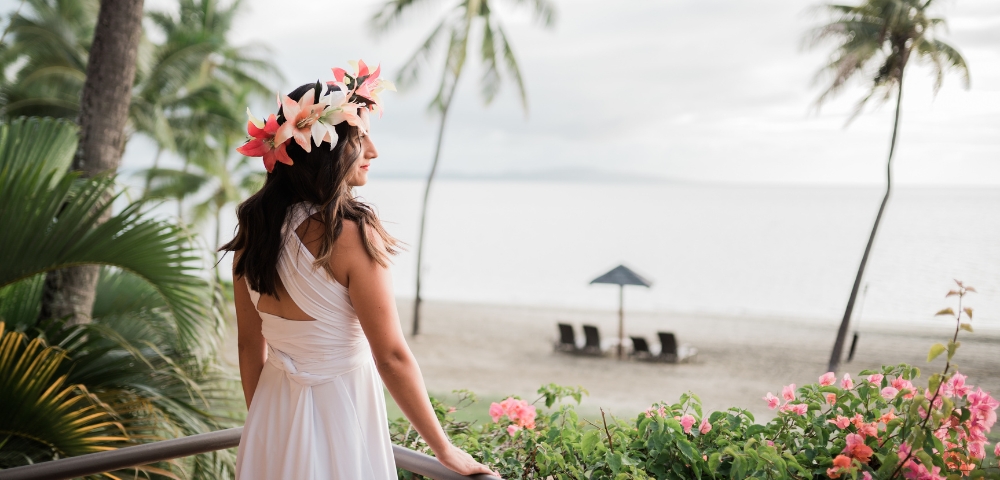 Woman in a flowing white dress and tropical flower crown gazes at a serene beach, framed by palm trees and pink blossoms in soft light.