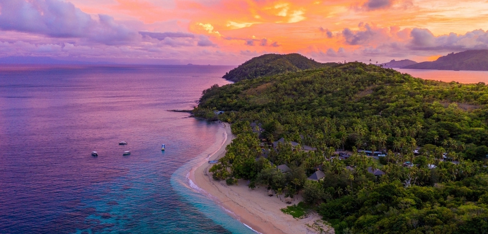 Aerial view of a lush tropical island, turquoise bay, sandy beach, forested hills, and boats beneath a vibrant pink-orange sunset sky.