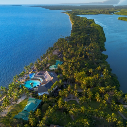 Aerial view of a tropical island resort with palm trees, turquoise pools, ocean and lagoon waterways, evoking a peaceful getaway.