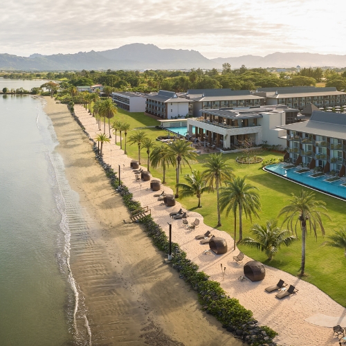 A modern beachfront resort features palm-lined sandy beaches, pools, green lawns, and mountains in the distance under a calm, bright sky.