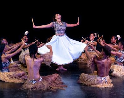 Dancers in traditional attire perform a rhythmic dance, with one dancer mid-air holding sticks, surrounded by seated dancers with sticks.