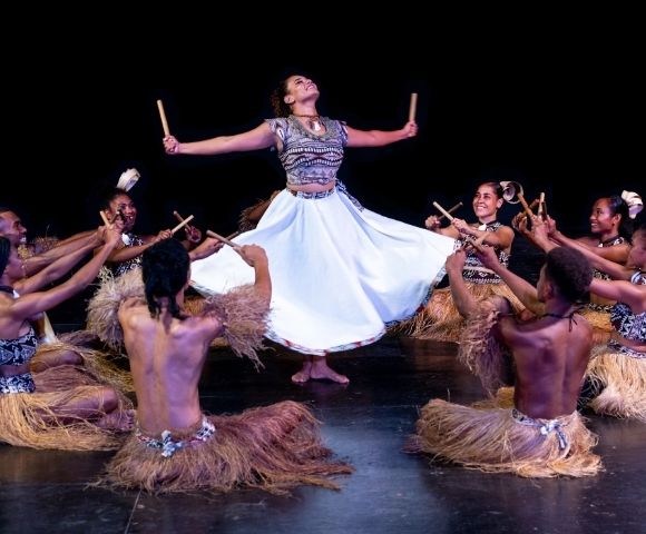 Dancers in traditional attire perform a rhythmic dance, with one dancer mid-air holding sticks, surrounded by seated dancers with sticks.