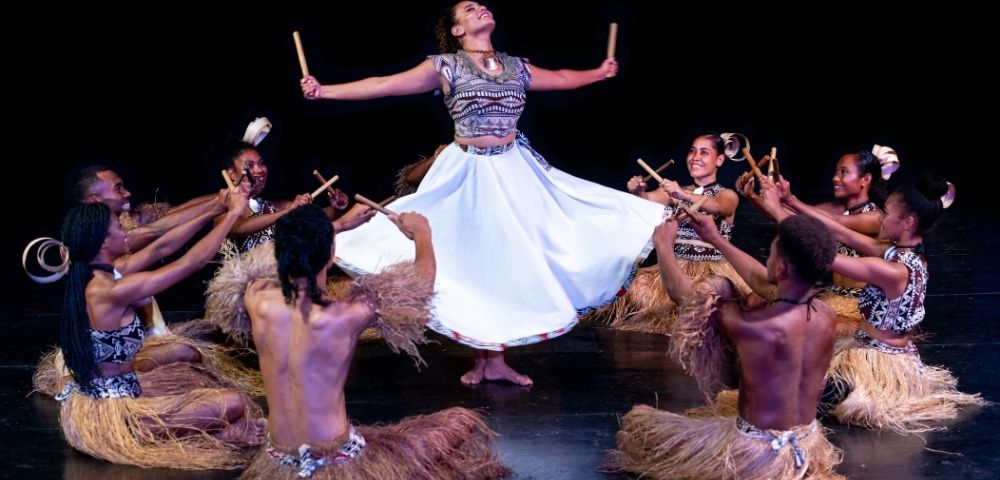 Dancers in traditional attire perform a rhythmic dance, with one dancer mid-air holding sticks, surrounded by seated dancers with sticks.