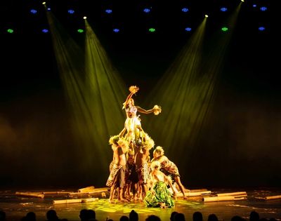 A group of performers in traditional attire forming a human pyramid on stage, illuminated by dramatic lighting.