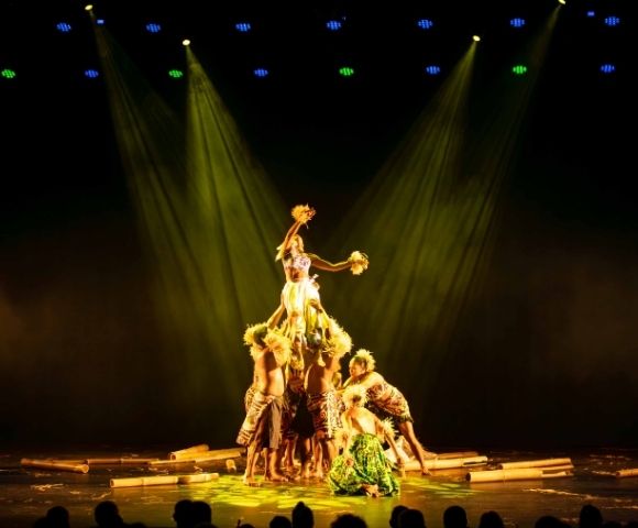 A group of performers in traditional attire forming a human pyramid on stage, illuminated by dramatic lighting.