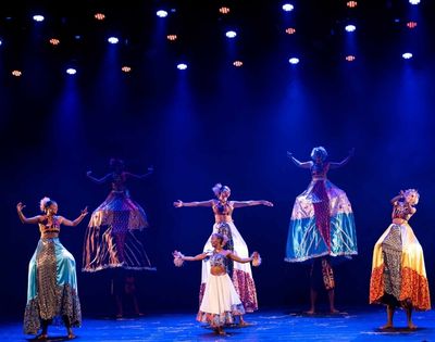 A group of performers dressed in colorful costumes dance under blue lights, some on stilts, showcasing traditional movement and expression.