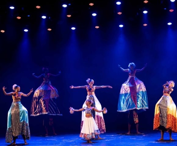 A group of performers dressed in colorful costumes dance under blue lights, some on stilts, showcasing traditional movement and expression.