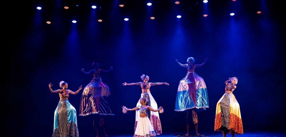 A group of performers dressed in colorful costumes dance under blue lights, some on stilts, showcasing traditional movement and expression.