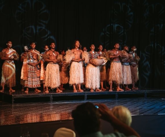 A group of performers dressed in traditional attire with grass skirts, standing on a stage, showcasing a cultural dance or performance.