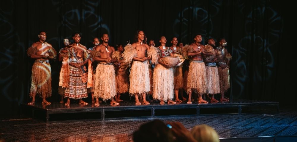 A group of performers dressed in traditional attire with grass skirts, standing on a stage, showcasing a cultural dance or performance.