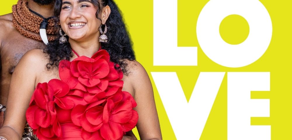 A couple dressed in traditional Fijian attire, with the woman wearing a red dress adorned with large flowers, smiling against a bright yellow background featuring the text 