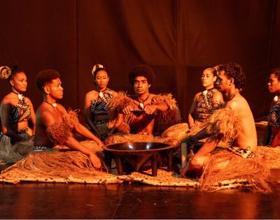 A group of performers in traditional attire sitting in a circle around a small fire on stage.